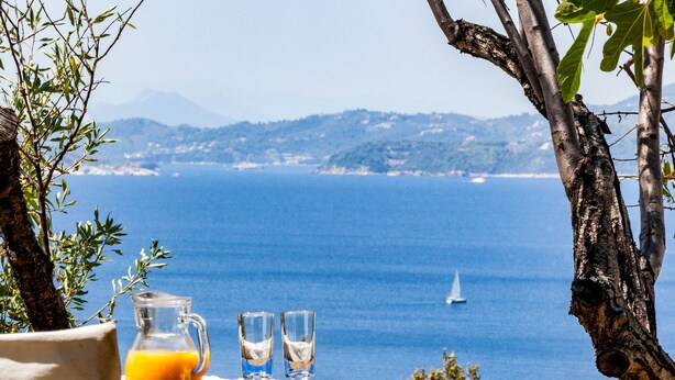 A table with a pitcher of orange juice, two glasses, and a plate with a book, overlooking a coastal view with a sailboat on the water.