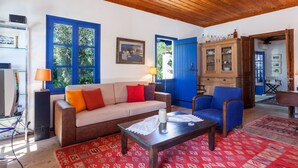 A cozy living room with a wooden ceiling, a sofa with red cushions, a blue armchair, a coffee table, and a cabinet with glassware.