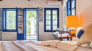 A bedroom with a bed, a desk, and a chair. There are open doors leading to a balcony with potted plants.