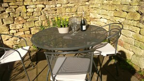 A round metal table with two chairs, a potted plant, a bottle of wine, and two glasses. - Wonderful Grade II Cotswold Stone Cottage in Perfect Location (Chipping Campden)
