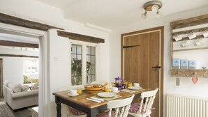 A dining area with a wooden table, white chairs, and a breakfast setting including a bowl of bread, cups, and a coffee maker. - Wonderful Grade II Cotswold Stone Cottage in Perfect Location (Chipping Campden)