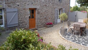 A stone house with a wooden door, a small garden with flowers and shrubs, and a patio area with a table and chairs.