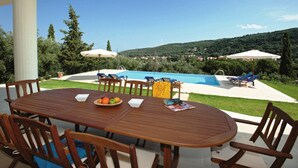 An outdoor dining area with a wooden table and chairs, a fruit bowl, and a view of a swimming pool and hills in the background. - Rent Villa In Corfu, Private Pool, Luxurious, Gorgeous Views, In House Catering (Klimatia Karousades)