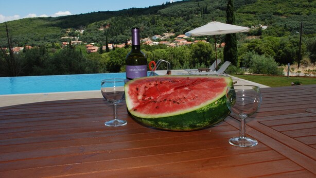 A wooden table with a watermelon, a bottle of wine, and two glasses, overlooking a pool and a hillside with houses. - Rent Villa In Corfu, Private Pool, Luxurious, Gorgeous Views, In House Catering (Klimatia Karousades)