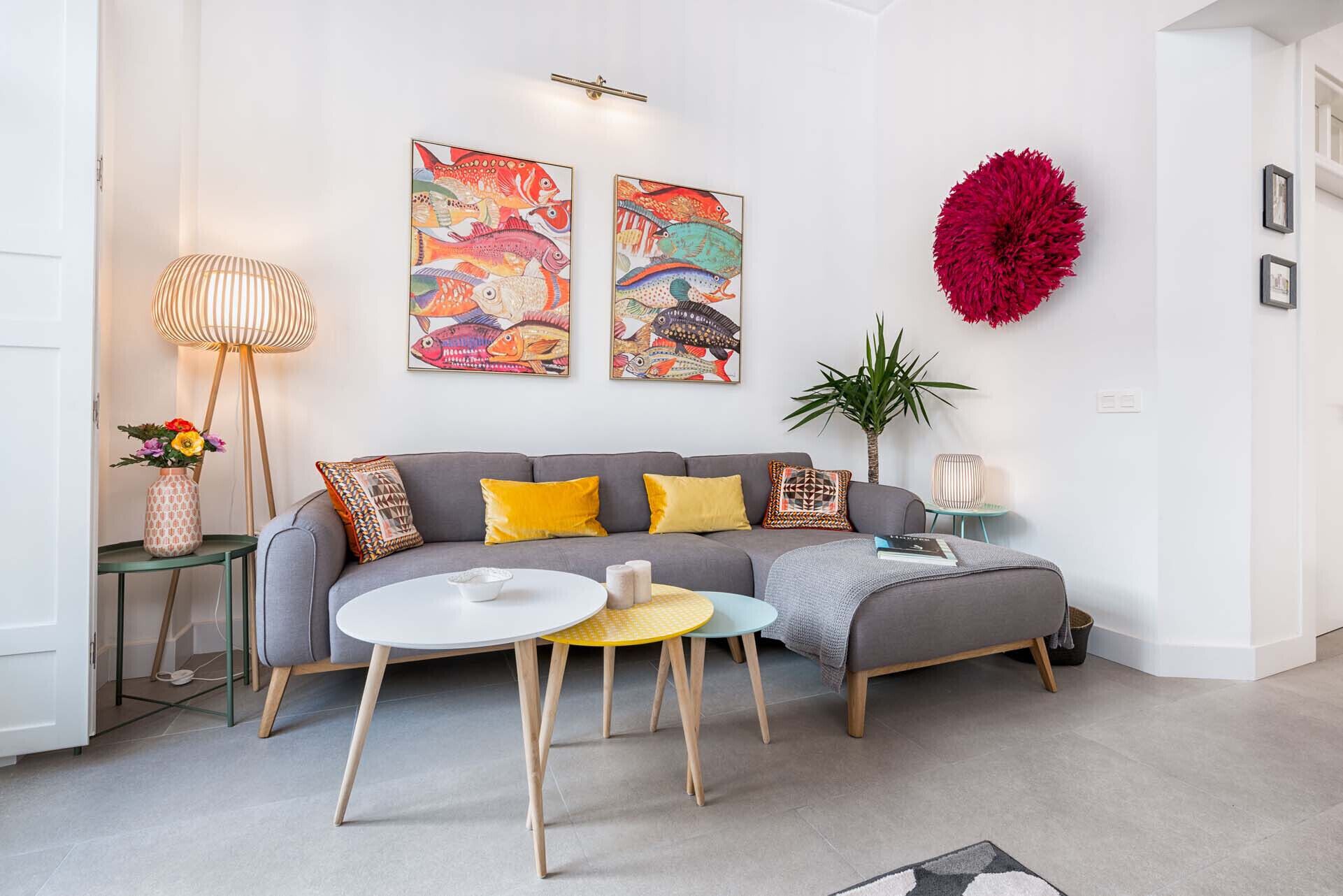 Un salon moderne avec un canapé gris, des coussins jaunes, une table basse ronde et deux tableaux colorés représentant des poissons, accrochés au mur.