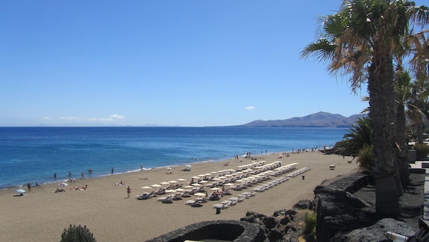 A beach with rows of sun loungers and umbrellas, a clear blue sky, and a mountain in the background.