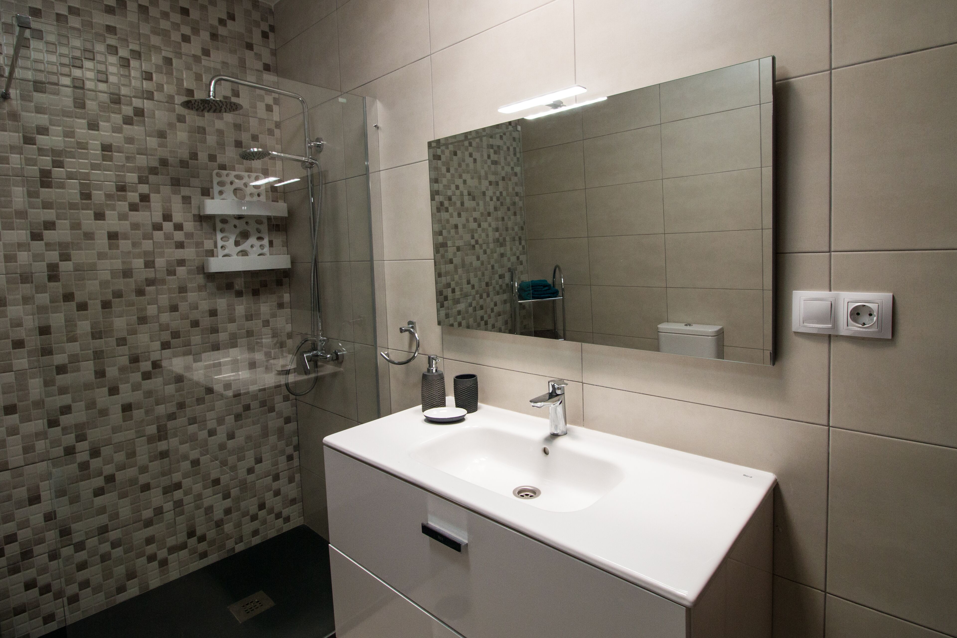 Ein modernes Badezimmer mit einem weißen Waschbecken, einer Duschkabine aus Glas und einer Wand mit Fliesenmosaik.