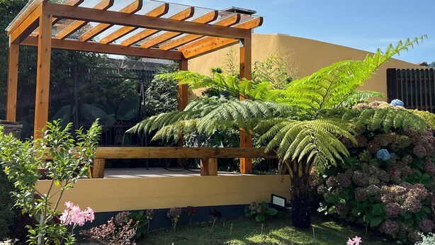 Una estructura de pérgola de madera con techo de vidrio, rodeada de exuberante vegetación y plantas en flor.