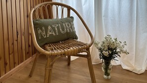 Una silla de mimbre con un cojín que dice 'NATURE' en una habitación con paneles de madera y una cortina blanca.