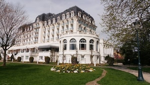 Un grand bâtiment blanc à plusieurs étages, doté d’une façade incurvée, entouré d’une pelouse bien entretenue et de parterres de fleurs.