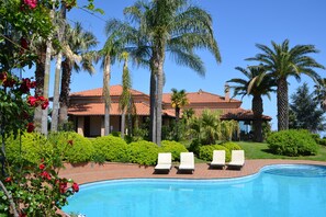 A pool area with lounge chairs, palm trees, and a building with a red-tiled roof.