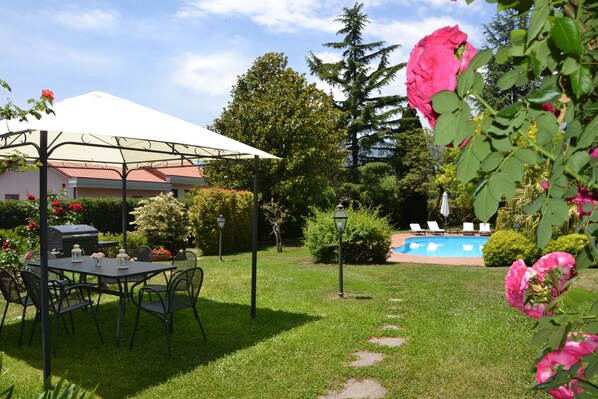 A covered outdoor seating area with a table and chairs, a pool, and a garden with various plants and flowers.