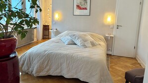 A bedroom with a large bed, bedside tables, a radiator, a potted plant, and a door. - SUPERB LOFT APARTMENT, CLASS 4*, SOUTH-FACING WITH TERRACE IN THE ORANGERIE DISTRICT (Strasbourg)
