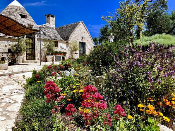 A stone building with a conical roof, surrounded by a garden with various flowers and plants. - Trullo With Private Pool (15x7), WI-FI free, tennis table,mountain bike for rent (Alberobello)