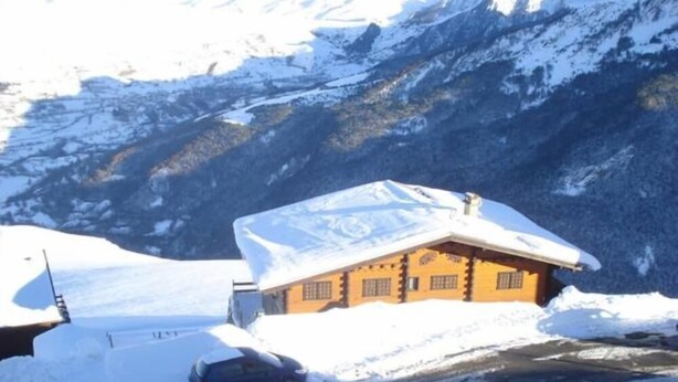 Une cabane en bois, dont le toit est recouvert de neige, entourée de neige et de montagnes.