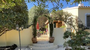 Un chemin de jardin menant à l’entrée d’un bâtiment, bordé de plantes en pot et entouré d’une zone dallée de pierres.