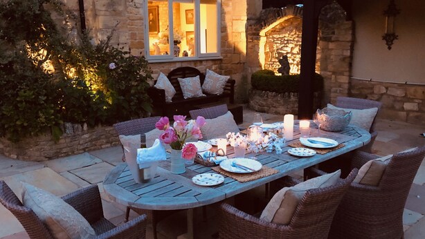 An outdoor dining area with a stone wall, a wooden table set for four, wicker chairs, and a bench with cushions. - Stunning Interior Designed 2 bedroom House in the center of Chipping Campden (Chipping Campden)