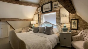 A bedroom with a stone wall, a bed with a white headboard, two bedside tables with lamps, and a chair. - Stunning Interior Designed 2 bedroom House in the center of Chipping Campden (Chipping Campden)
