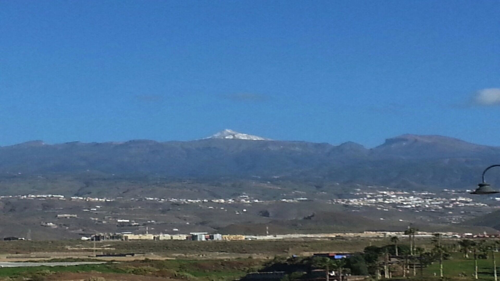 Een berg met een besneeuwde top op de achtergrond, een stad met gebouwen in de voorgrond, en een heldere blauwe lucht.