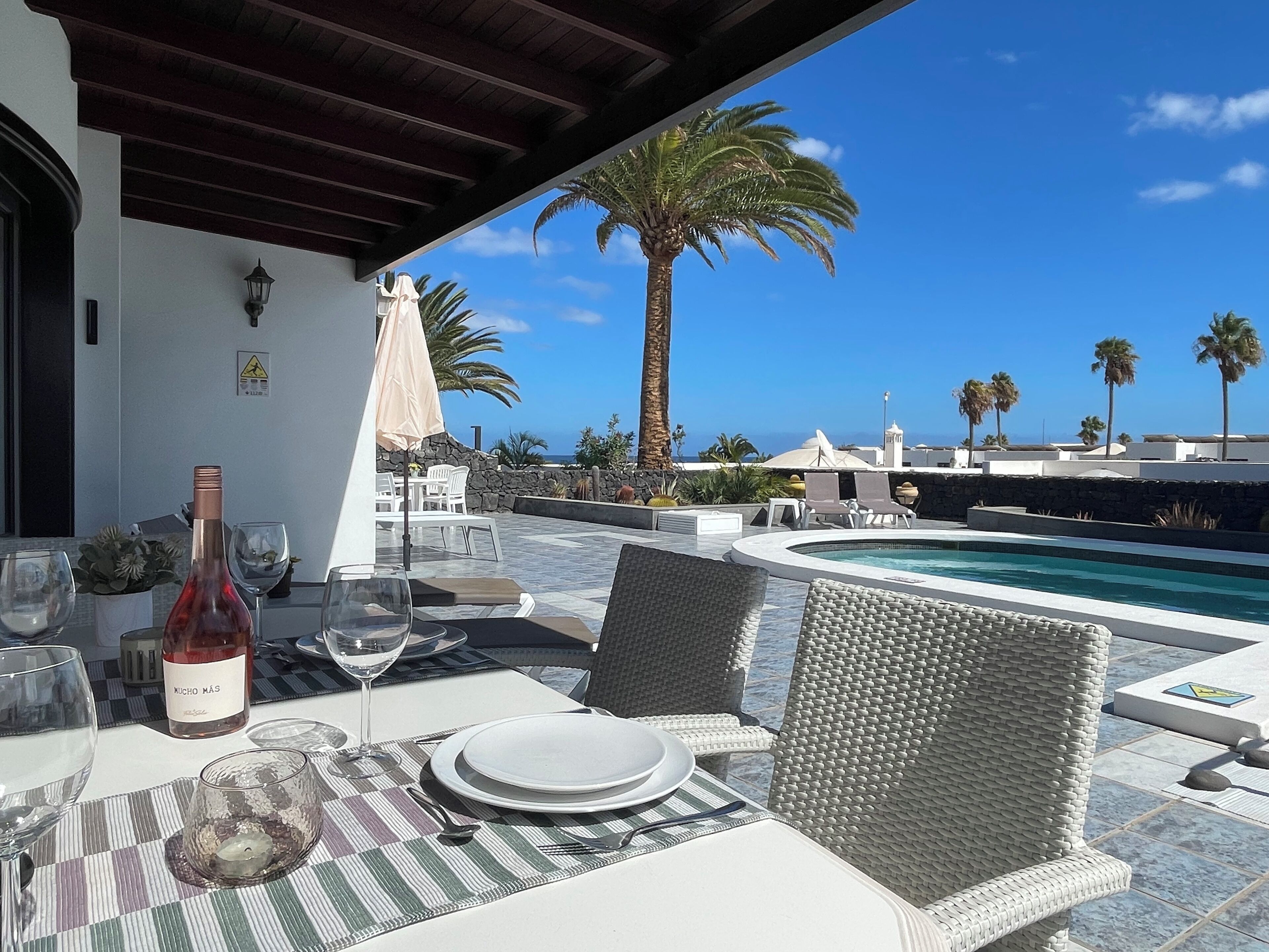 A dining area with a table set for two, a bottle of wine, and a glass of water. There is a pool in the background with palm trees and a clear blue sky.