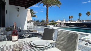 A dining area with a table set for two, a bottle of wine, and a glass of water. There is a pool in the background with palm trees and a clear blue sky.