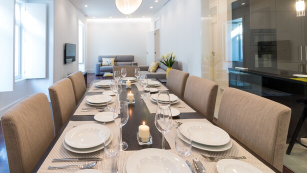 A modern dining room with a long table set for a meal, a sofa in the background, and a kitchen area visible through a glass partition. - House Sweet Home - Lisbon Center (Lisboa)