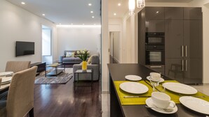 A modern living room with a sofa, coffee table, and a flat-screen TV. There is a dining area with a table set for two, and a kitchen with dark cabinets and stainless steel appliances. - House Sweet Home - Lisbon Center (Lisboa)