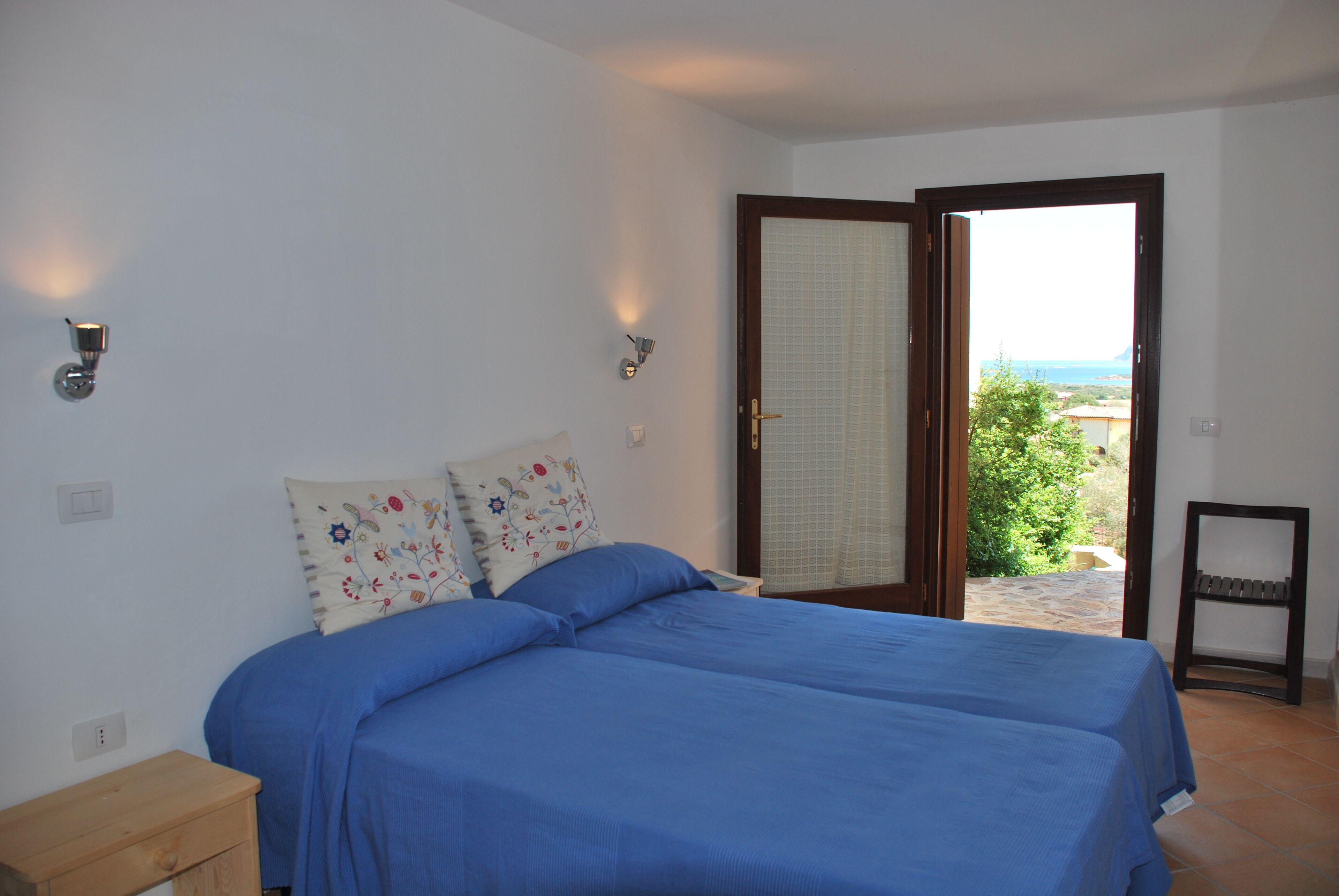 Un dormitorio con cama, dos almohadas, una mesita de noche y vistas al exterior a través de una puerta abierta.
