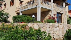 A two-story house with a stone facade, a balcony with a metal railing, and a small garden with various plants and flowers.