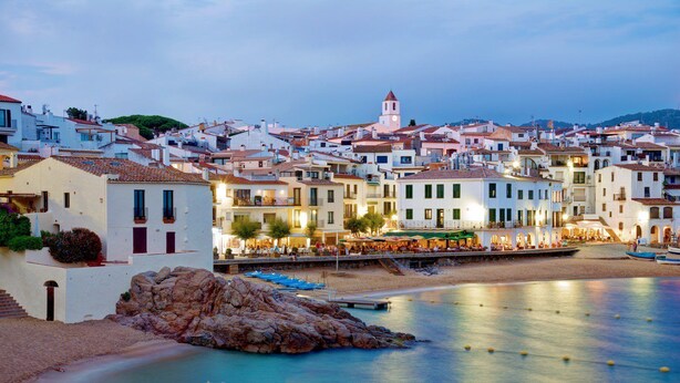 Un pueblo costero con edificios blancos, una torre destacada y un frente marítimo tranquilo con barcos.