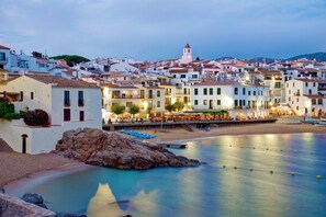 A coastal town with white buildings, a prominent tower, and a calm waterfront with boats.