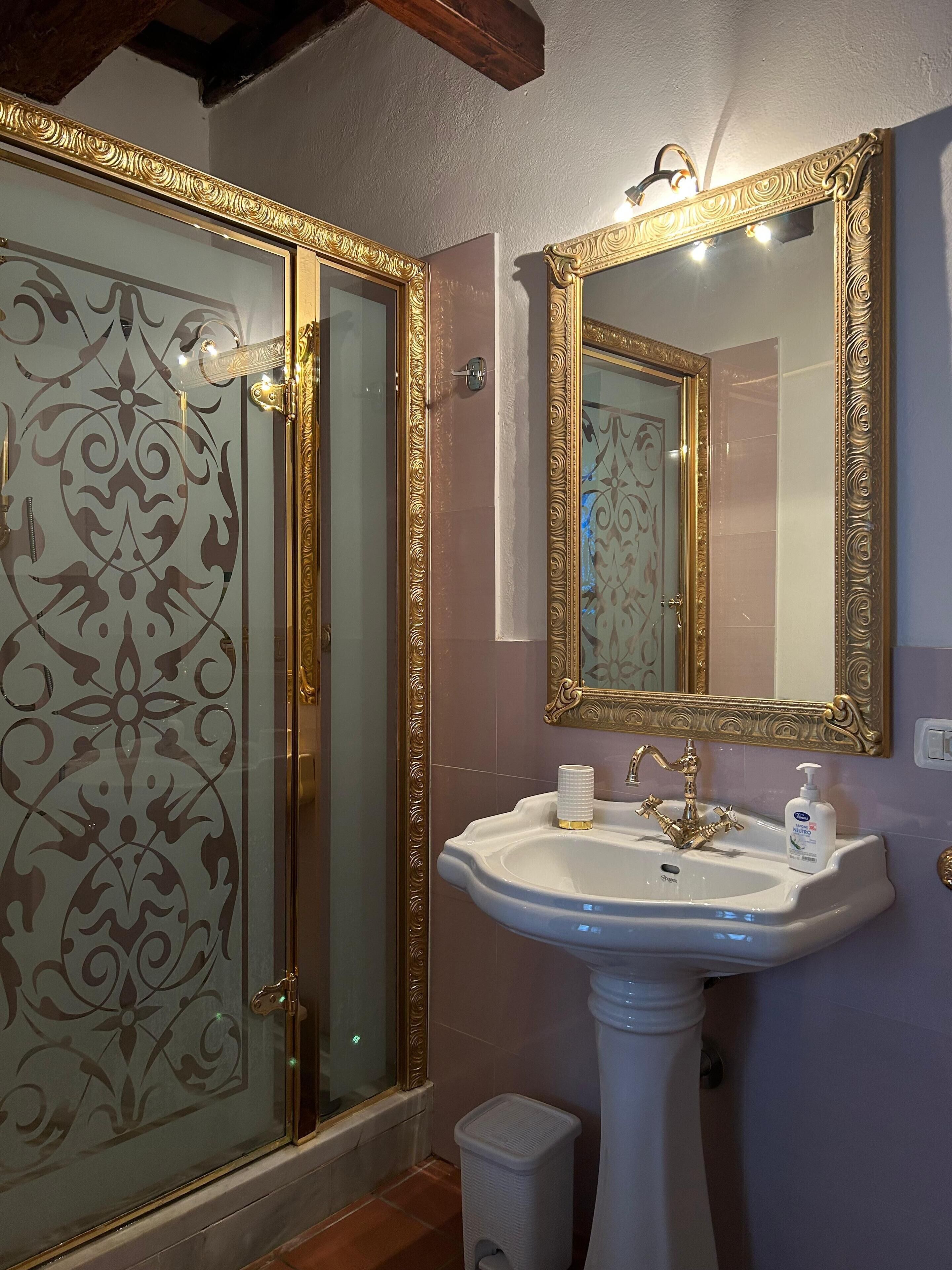 Un baño con lavamanos blanco, espejo con marco dorado y cabina de ducha de vidrio con un diseño decorativo.