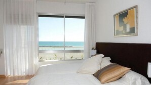 A bedroom with a large bed, white bedding, a dark headboard, a painting on the wall, and a view of the sea through a window with sheer curtains. - Luxury beachside villa  with private pool, terrace and sea views. (Benicassim)
