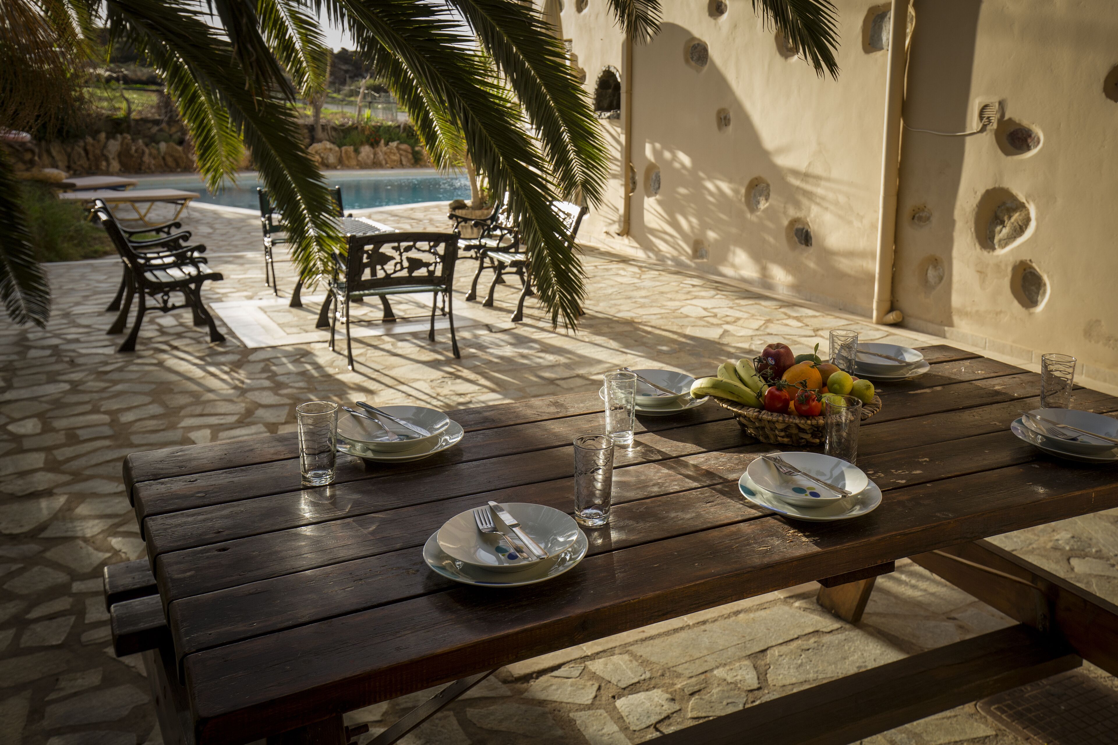 A wooden table set for dining with plates, cutlery, and glasses, a fruit basket, and a view of a swimming pool and palm trees in the background.