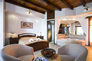 Habitación de hotel con cama, dos sillas, una mesa de comedor con un cuenco de fruta, un cuadro en la pared, un baño con bañera y un espejo.