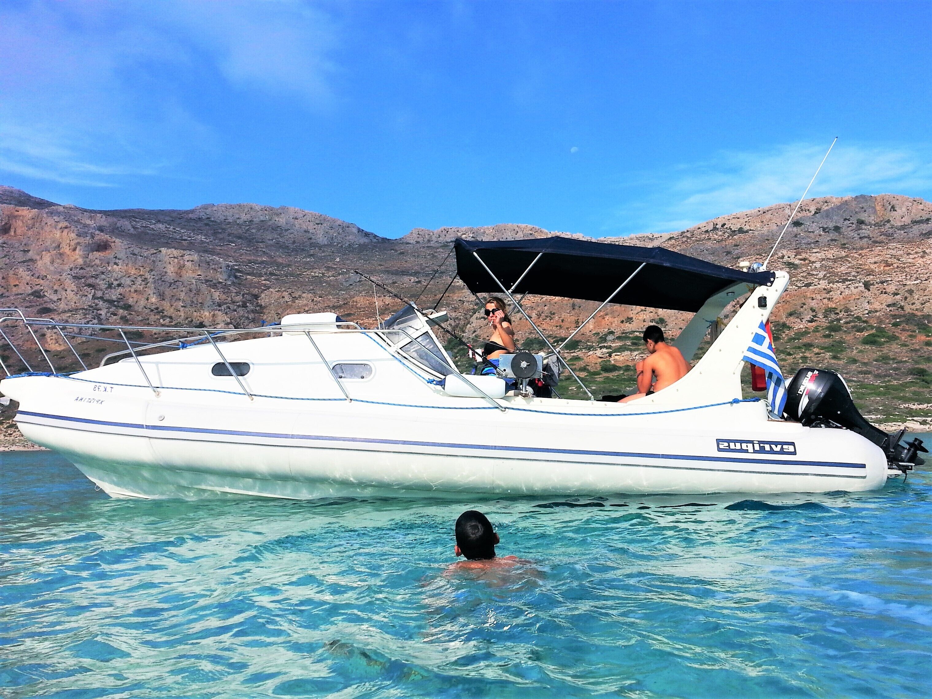 Un bateau avec une cabine, une personne qui nage et un paysage rocheux en arrière-plan.