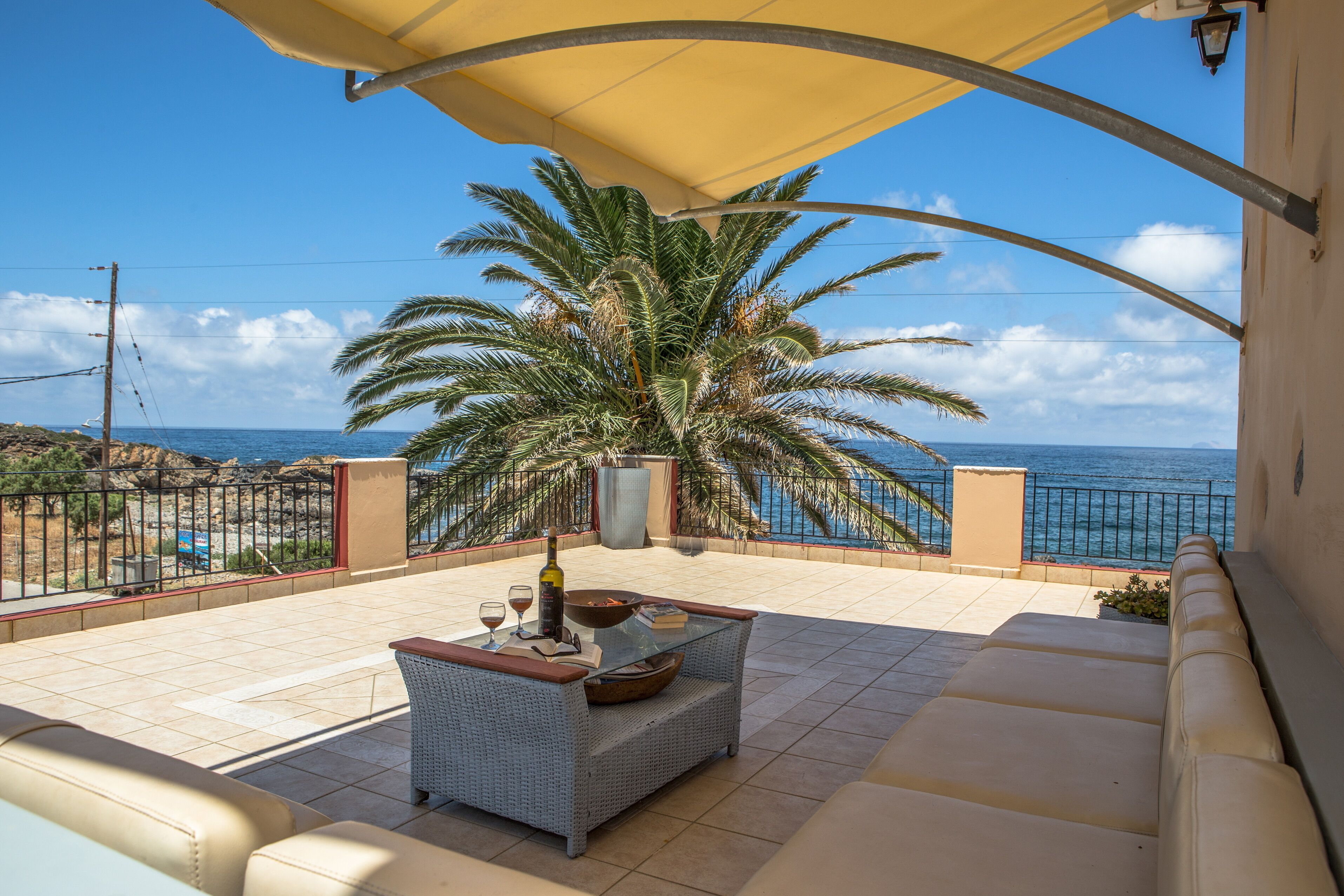 A balcony with a view of the sea, a wicker sofa, a table with wine glasses, and a palm tree.