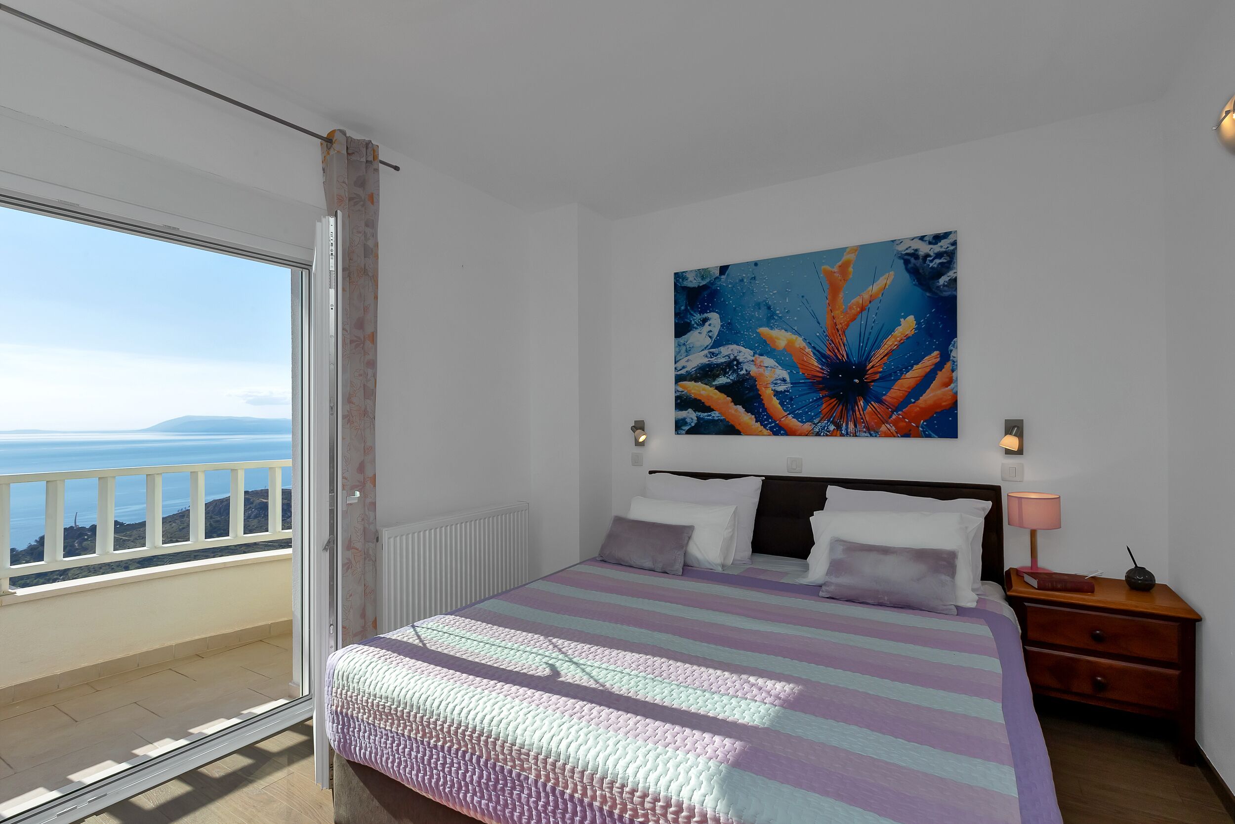 A bedroom with a large bed, a nightstand, a painting, and a view of the sea.