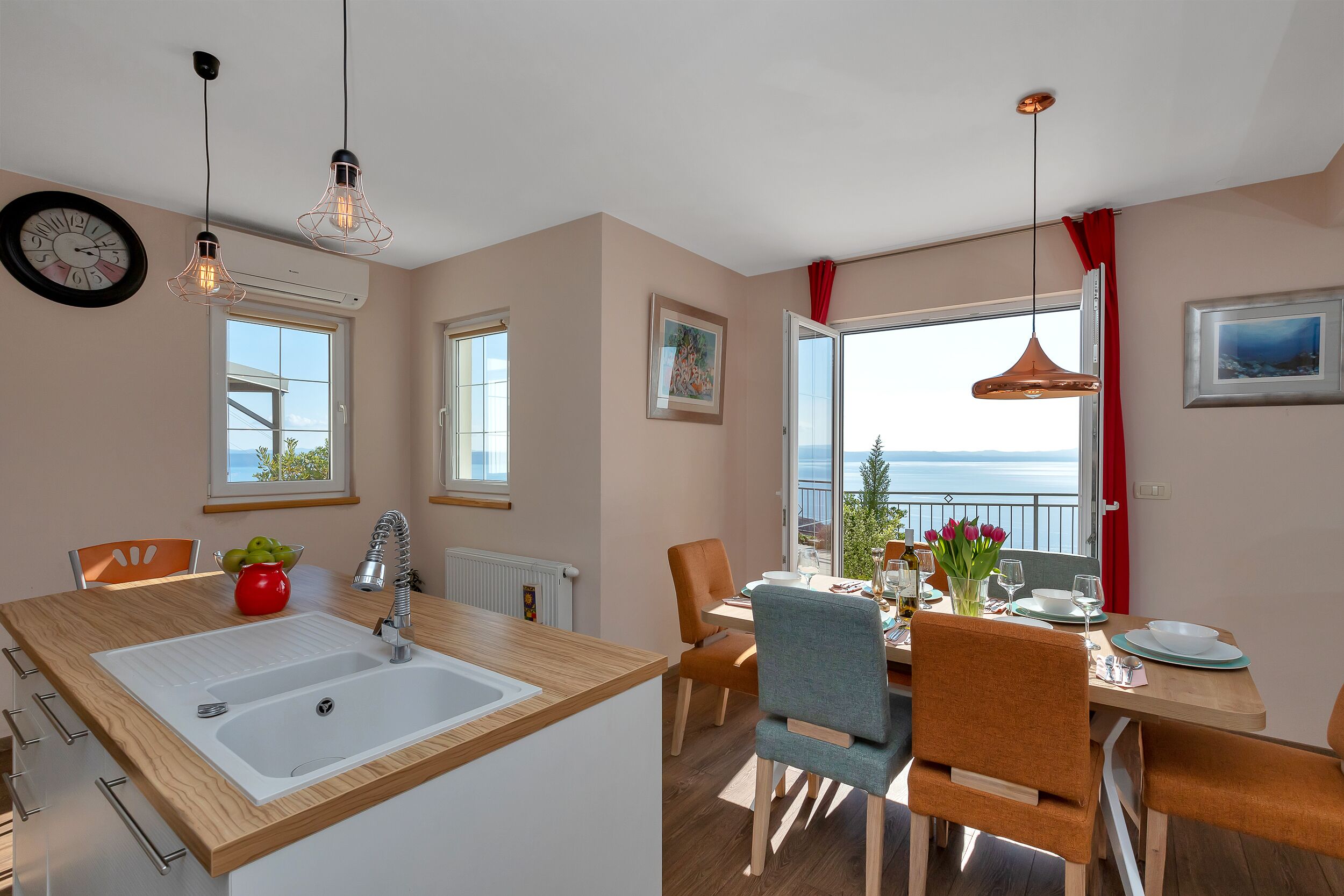 A modern kitchen with a large island, a dining area with a view, and a balcony with red curtains.