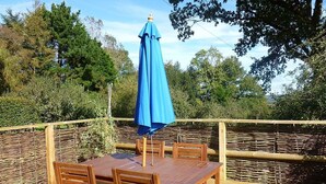 A wooden deck with a table and chairs, a blue umbrella, and a wicker fence. - Rural Holiday Cottage with Wonderful Countryside Views on Friendly Smallholding. (Stockland)