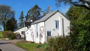 A white two-story house with a sloped roof, a small porch, and a garden with flowering plants. - Rural Holiday Cottage with Wonderful Countryside Views on Friendly Smallholding. (Stockland)