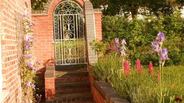 Un bâtiment en briques, agrémenté d’une grille en métal décorative et d’un escalier menant à un jardin où poussent différentes variétés de fleurs.