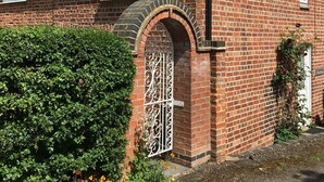 Une maison en briques avec un toit en tuiles, une porte blanche ornée d’un portail en métal décoratif, et un petit jardin avec des fleurs et une haie.