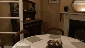 Une salle à manger avec une table ronde recouverte d’une nappe à motifs floraux, entourée de chaises. On y trouve une cheminée surmontée d’un miroir, ainsi qu’un tableau encadré accroché au mur.