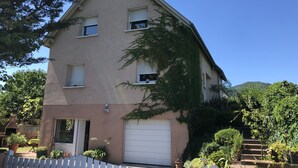 A two-story house with a white garage door, a small porch with a white picket fence, and a garden with various plants and a potted tree. - Apartment/ flat - Wettolsheim (Wettolsheim)