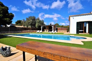 Une piscine avec une terrasse en bois, un espace extérieur couvert aménagé en salon, et un bâtiment blanc doté d’un toit en tuiles.