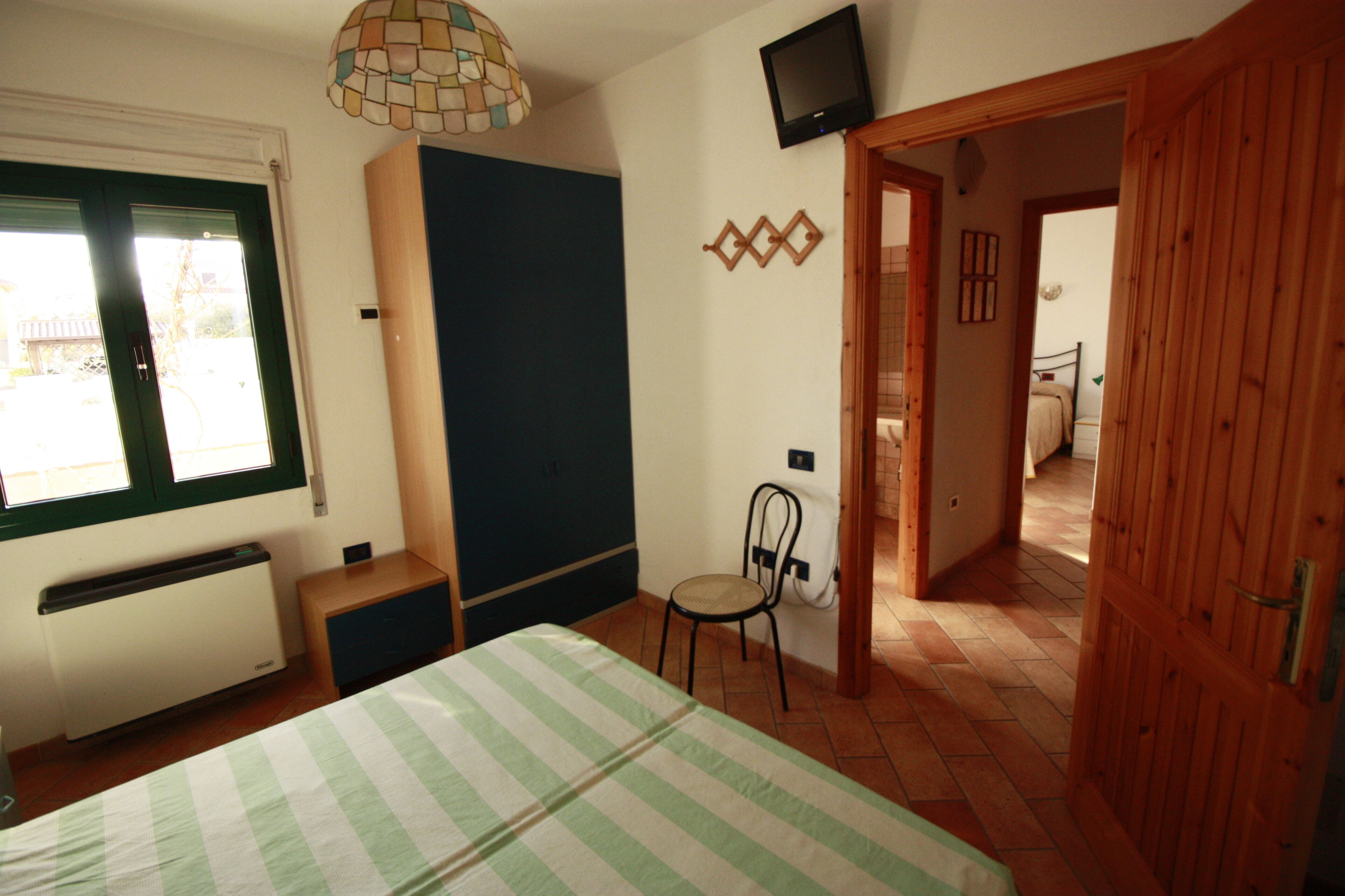 Un dormitorio con cama, sillón, radiador, televisor y puerta que da a otra habitación.