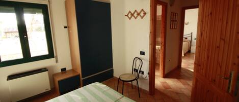 Un dormitorio con cama, sillón, radiador, televisor y puerta que da a otra habitación.