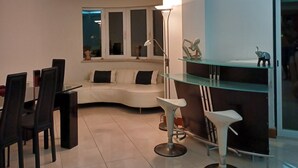 Una sala de estar moderna con un sofá blanco curvo, una mesa de comedor con sillas, una lámpara de pie y una mesa de centro de vidrio.