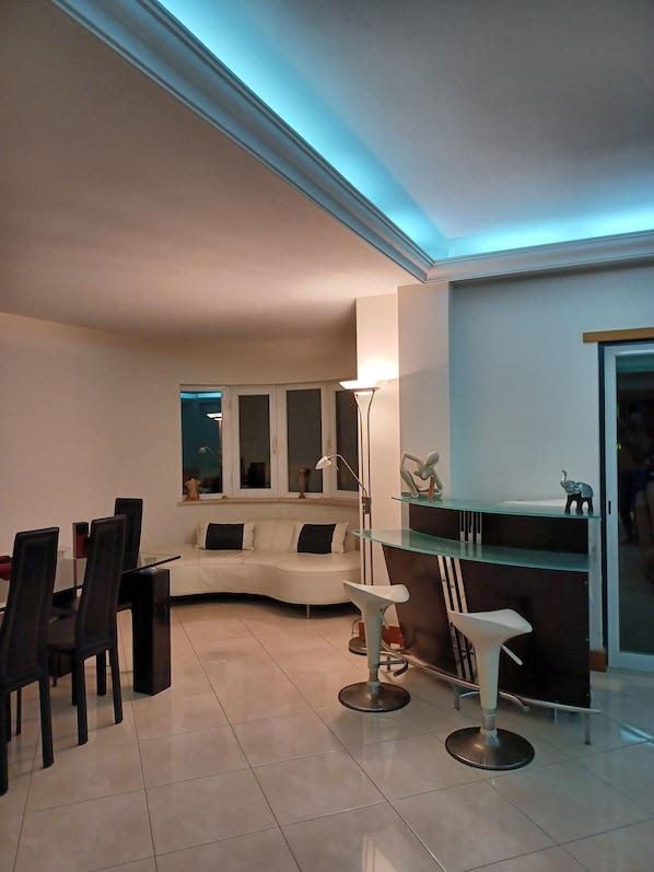 Una sala de estar moderna con un sofá blanco curvo, una mesa de comedor con sillas, una lámpara de pie y una mesa de centro de vidrio.